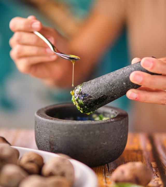 Soft focus of crop female adding spoonful of olive oil to mortar with mojo de cilantro sauce while cooking papas de Tenerife dish for lunch